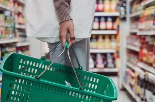 Side view of a woman shopping in a supermarket aisle holding a green basket and a drink bottle.
