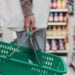 Side view of a woman shopping in a supermarket aisle holding a green basket and a drink bottle.